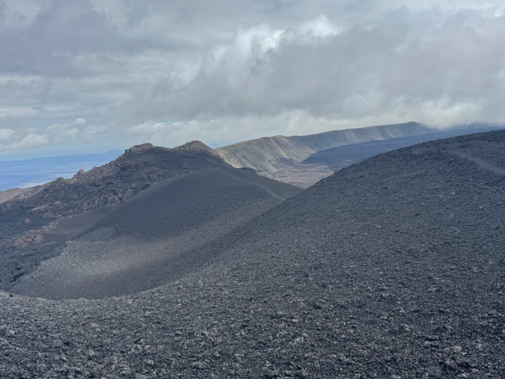 An S-curve of black cinders fills the foreground. Beyond, the curve of a volcanic crater is visible. The distant curve is green with vegetation, but the closer slopes are black and lifeless.