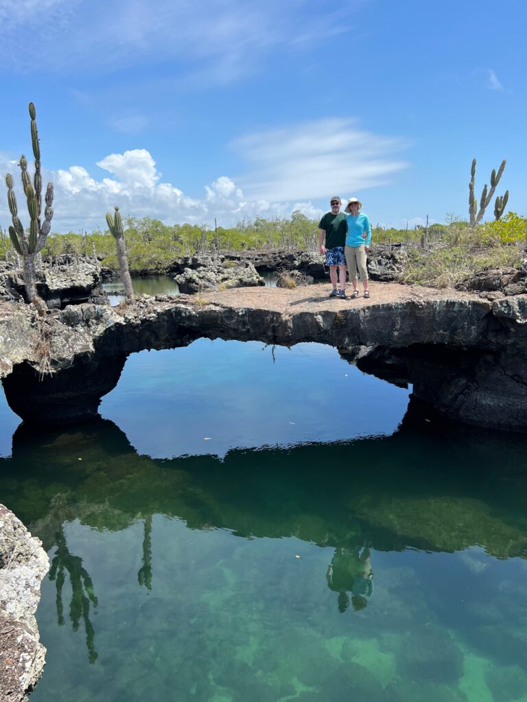 Two people stand on an arch of volcanic stone over the water. A large cactus is visible on the left, and there is a layer of green mangroves in the distance.